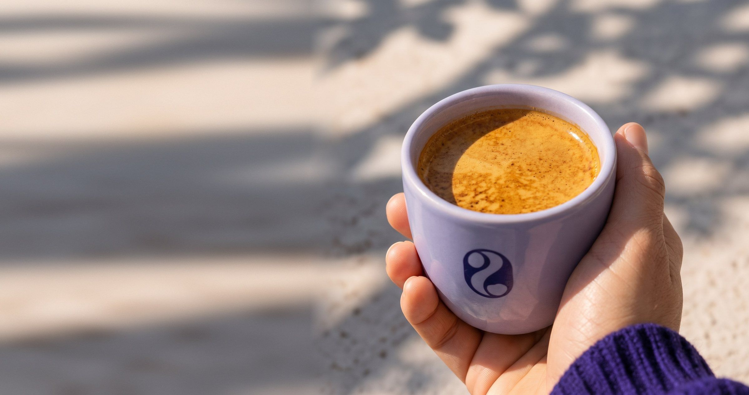 Hand houdt een lila espressokopje met het easybeans-beeldmerk vast, in zonlicht met bladschaduw